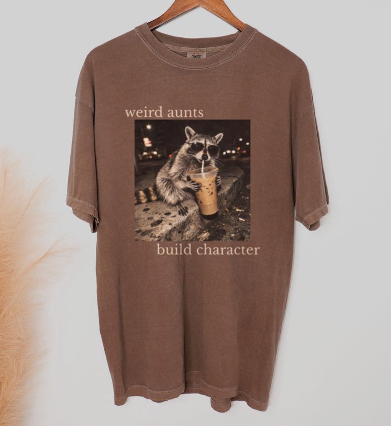 May include: Brown t-shirt with the words "weird aunts build character" printed on it. The shirt features a graphic of a raccoon wearing sunglasses and drinking an iced coffee.