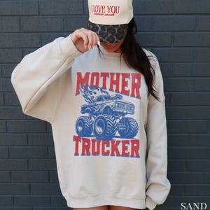 May include: Sand-colored sweatshirt with a graphic of a blue monster truck with flames and the words "MOTHER TRUCKER" in red. The sweatshirt is paired with a cream-colored baseball cap that says "I LOVE YOU" in red.