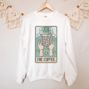 Puede incluir: Sudadera blanca con un dise&ntilde;o de carta del tarot que representa una taza de caf&eacute;. El dise&ntilde;o incluye manos, estrellas y el texto "THE COFFEE". La sudadera est&aacute; colgada en una percha de madera.