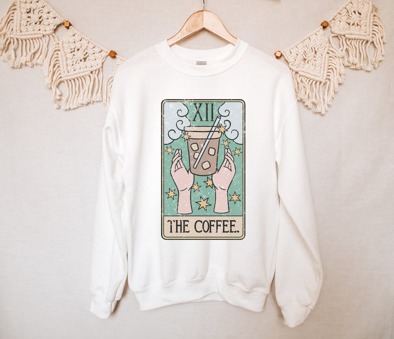 Puede incluir: Sudadera blanca con un dise&ntilde;o de carta del tarot que representa una taza de caf&eacute;. El dise&ntilde;o incluye manos, estrellas y el texto "THE COFFEE". La sudadera est&aacute; colgada en una percha de madera.