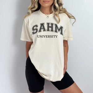 Puede incluir: Una camiseta de color crema con texto negro que dice "SAHM UNIVERSITY".