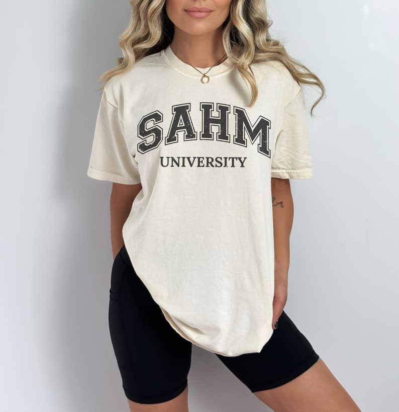 Puede incluir: Una camiseta de color crema con texto negro que dice "SAHM UNIVERSITY".