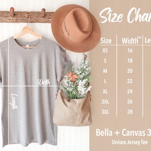 May include: Size chart for a unisex jersey tee, showing measurements in inches for width and length. The chart includes sizes XS through 3XL. The image also shows a grey t-shirt hanging on a wooden hanger, a brown hat, and a brown bag.
