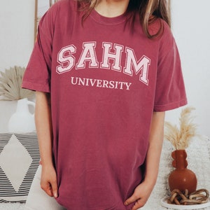 Puede incluir: Una camiseta de gran tama&ntilde;o de color marr&oacute;n con el texto "SAHM UNIVERSITY" impreso en blanco.
