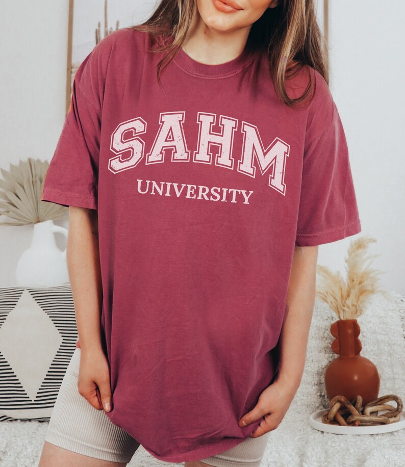Puede incluir: Una camiseta de gran tama&ntilde;o de color marr&oacute;n con el texto "SAHM UNIVERSITY" impreso en blanco.