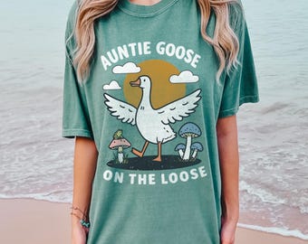 Może przedstawiać: Jasnozielony t-shirt z kreskówkową gęsią i grzybami. Tekst na koszulce brzmi "Auntie Goose on the Loose".