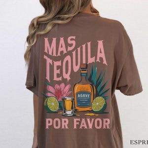 May include: Brown t-shirt with a pink graphic of a tequila bottle, limes, and a shot glass. The text on the shirt reads "Mas Tequila Por Favor".