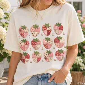 Puede incluir: Una camiseta de manga corta color marfil con un estampado gráfico de doce fresas dispuestas en cuadrícula. Cada fresa está compuesta por varios patrones de patchwork en tonos rojos, rosas y blancos, con hojas verdes de estilo bordado.