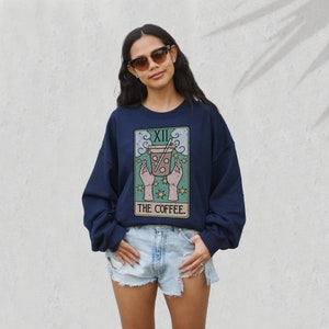 Puede incluir: Sudadera azul marino con dise&ntilde;o de carta del tarot que representa una taza de caf&eacute;. La carta dice "XII" en la parte superior y "THE COFFEE" en la inferior. El dise&ntilde;o incluye manos, estrellas y una taza de caf&eacute; con pajita.