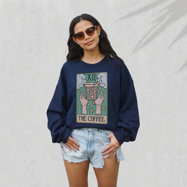 Puede incluir: Sudadera azul marino con dise&ntilde;o de carta del tarot que representa una taza de caf&eacute;. La carta dice "XII" en la parte superior y "THE COFFEE" en la inferior. El dise&ntilde;o incluye manos, estrellas y una taza de caf&eacute; con pajita.