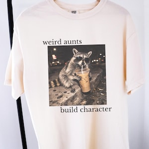 May include: A cream-coloured t-shirt with a raccoon in sunglasses drinking from a cup. The text "weird aunts" is above the image, and "build character" is below. The shirt is hanging on a black clothing rack.