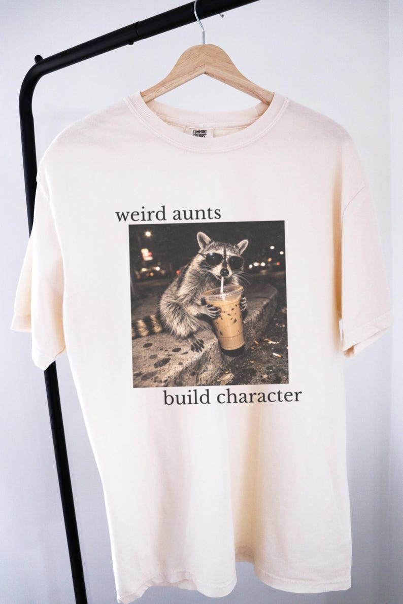 May include: A cream-coloured t-shirt with a raccoon in sunglasses drinking from a cup. The text "weird aunts" is above the image, and "build character" is below. The shirt is hanging on a black clothing rack.
