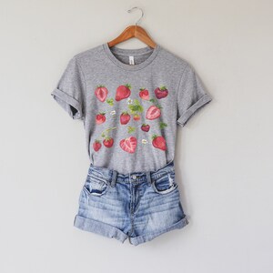 May include: A grey t-shirt with a graphic print of red strawberries and white flowers. The shirt is paired with light blue denim shorts.