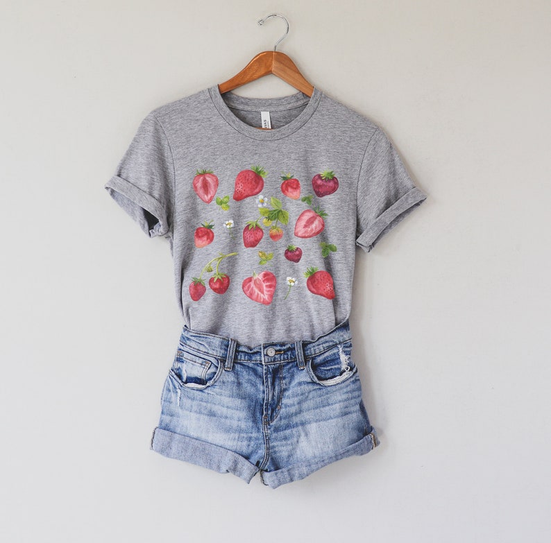 May include: A grey t-shirt with a graphic print of red strawberries and white flowers. The shirt is paired with light blue denim shorts.