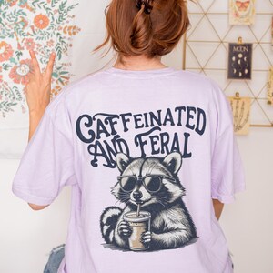 May include: A lavender t-shirt with a graphic of a raccoon wearing sunglasses and drinking a beverage. The text "caffeinated and feral" is printed on the shirt.