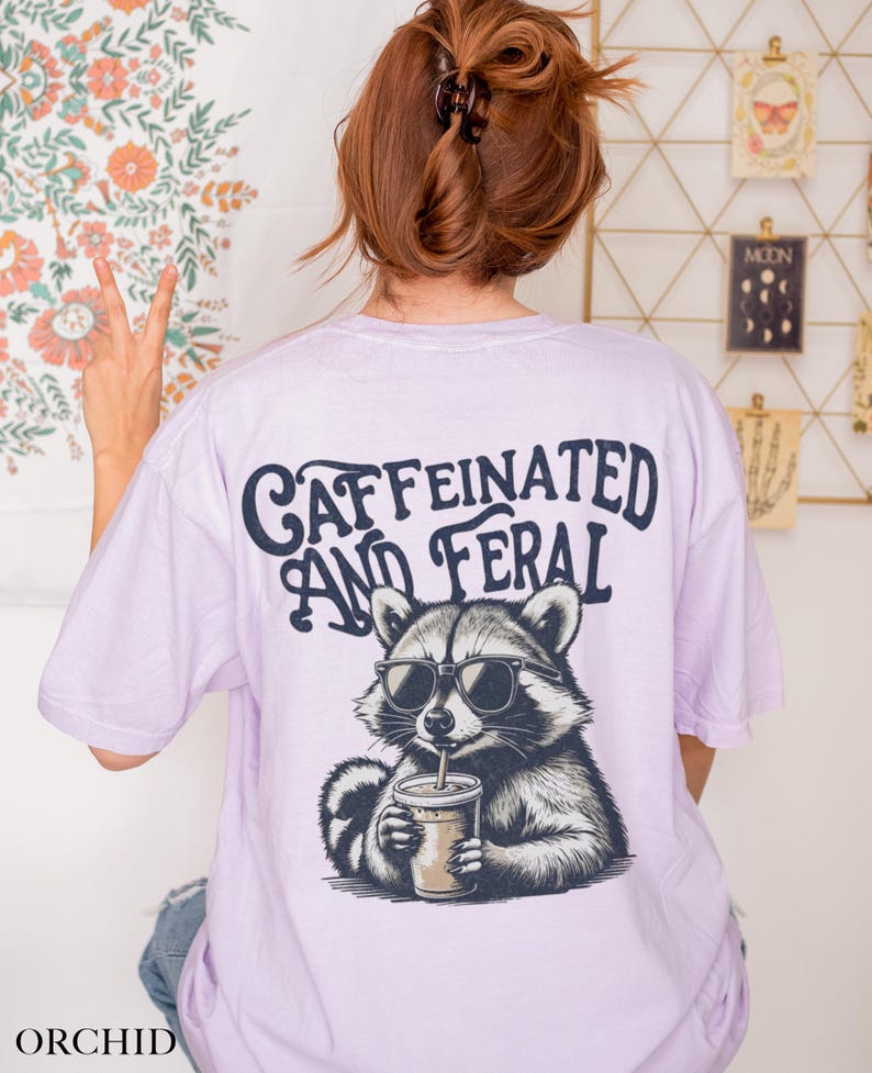 May include: A lavender t-shirt with a graphic of a raccoon wearing sunglasses and drinking a beverage. The text "caffeinated and feral" is printed on the shirt.