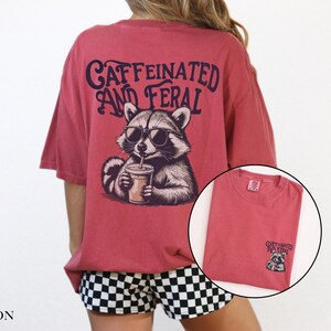 May include: A red t-shirt with a graphic of a raccoon wearing sunglasses and holding a cup of coffee. The text "CAFFEINATED AND FERAL" is printed on the shirt.