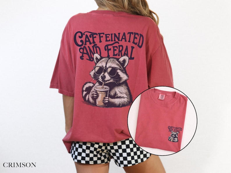 May include: A red t-shirt with a graphic of a raccoon wearing sunglasses and holding a cup of coffee. The text "CAFFEINATED AND FERAL" is printed on the shirt.