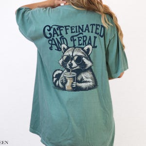 May include: A light green t-shirt with a graphic of a raccoon wearing sunglasses and drinking a beverage. The text "caffeinated and feral" is printed on the shirt.
