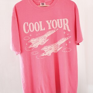 Puede incluir: Una camiseta rosa con las palabras "COOL YOUR" en blanco roto, sobre dos ilustraciones estilizadas de aviones a reacci&oacute;n. La camiseta est&aacute; colgada de una percha de madera.