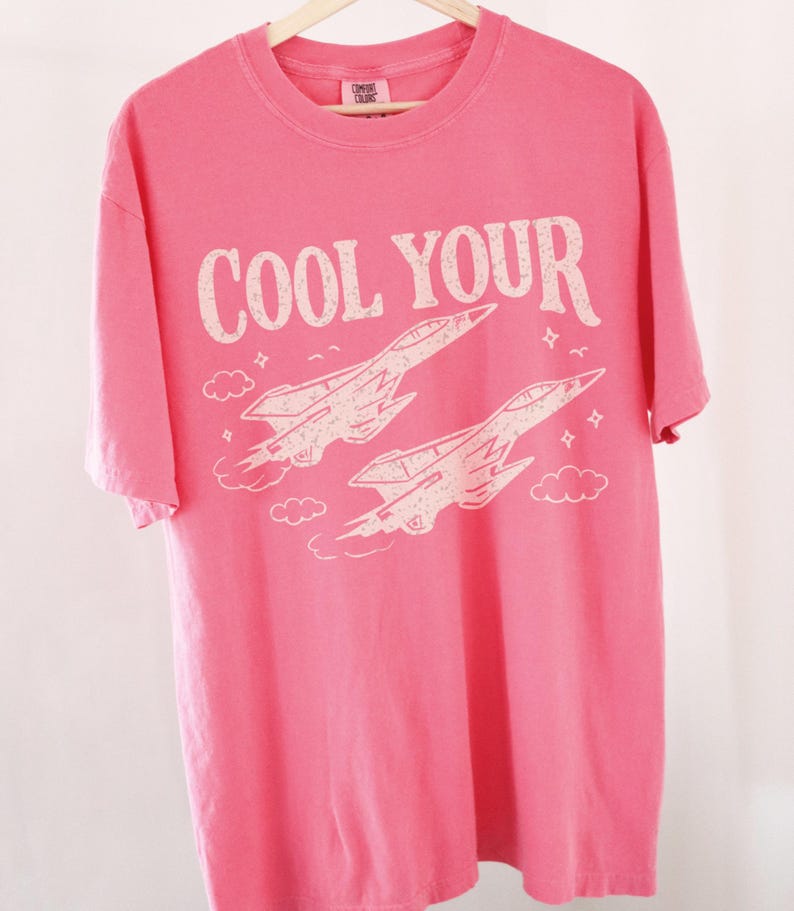 Puede incluir: Una camiseta rosa con las palabras "COOL YOUR" en blanco roto, sobre dos ilustraciones estilizadas de aviones a reacci&oacute;n. La camiseta est&aacute; colgada de una percha de madera.