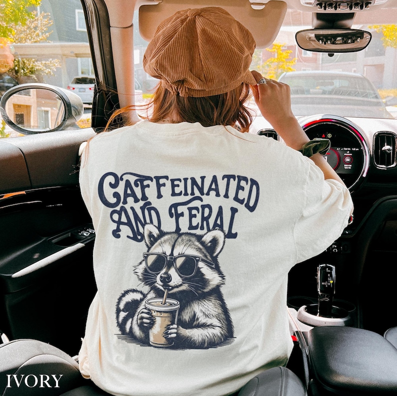 May include: A white t-shirt with a graphic of a raccoon wearing sunglasses and drinking a beverage. The text "CAFFEINATED AND FERAL" is printed on the shirt.
