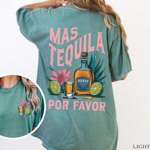 May include: Light green t-shirt with a pink graphic of a tequila bottle, limes, and a shot glass. The text on the shirt reads "Mas Tequila Por Favor".