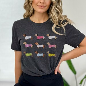 May include: A dark gray t-shirt with a graphic print of nine dachshunds wearing different patterned pajamas. The dachshunds are all facing the right side of the image.