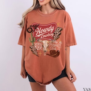 May include: Burnt orange t-shirt with a graphic design featuring the text "Howdy Cowboy" and "The Cowgirl Club". The design includes a steer skull, cactus, lightning bolts, a disco ball, and a cowboy hat.