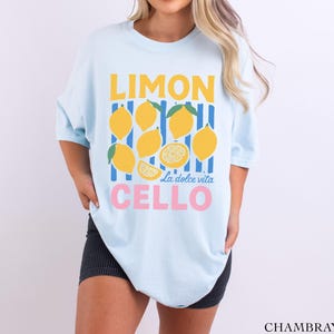 May include: Light blue t-shirt with a lemon-themed graphic. The design features yellow lemons, blue and yellow stripes, and the words "LIMON CELLO" in yellow and pink, with "La dolce vita" in the middle.