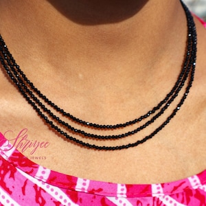 May include: A close-up of a black beaded necklace with three strands. The necklace is worn against a pink and white patterned top. The jewelry is delicate and elegant, suitable for various occasions.