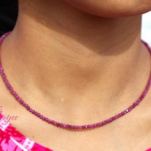 May include: A fine, beaded necklace in a deep magenta colour. The necklace is made up of small, evenly sized beads, forming a continuous strand. The necklace is worn against a pink and white patterned top.