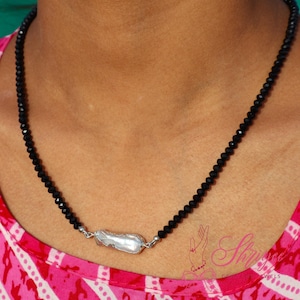 Könnte beinhalten: Eine schwarze Perlenkette mit einem großen, unregelmäßig geformten, weißen Anhänger. Die Kette wird über einem rosa-weiß gemusterten Oberteil getragen. Der Anhänger ist mit silberfarbenem Metall befestigt.