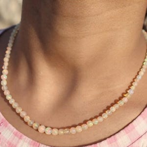 Puede incluir: Un collar compuesto por pequeñas cuentas de ópalo redondas e iridiscentes que muestran sutiles destellos de color. Las cuentas están ensartadas en una sola hebra que descansa alrededor del cuello. El texto "Shreejee GEMS AND JEWELS" es visible.