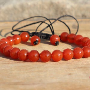 Puede incluir: Una pulsera de cuentas rojas con un cordón negro ajustable. La pulsera presenta cuentas redondas y pulidas. El fondo es una superficie de madera marrón claro. Es una joya.