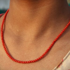 Op de afbeelding: Een felrode kralenketting om de nek van een persoon. De ketting bestaat uit kleine, ronde kralen. De persoon draagt een top met bloemenpatroon in rood en wit.