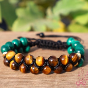 May include: A handcrafted bracelet featuring alternating rows of green malachite and tiger's eye beads. The beads are strung on a black woven cord. The bracelet is displayed on a wooden surface with a blurred green background.