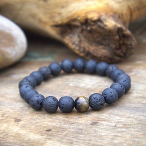 May include: A dark grey lava stone bracelet with a single, round, brown tiger eye bead. The bracelet is strung on an elastic band. The beads are approximately 1 cm in diameter. The bracelet is displayed on a wooden surface.