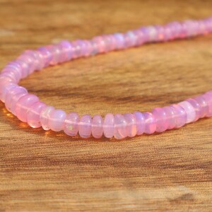 May include: A strand of pink opal beads on a wooden surface. The beads are small and round, and they have a subtle iridescent sheen.