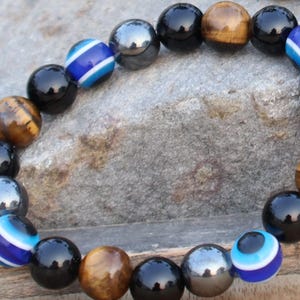 May include: A beaded bracelet with black, brown, and gray beads. The bracelet also features blue and white evil eye beads.