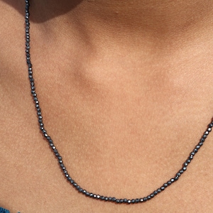 Könnte beinhalten: Eine zarte schwarze Perlenkette aus kleinen, facettierten Perlen hängt an einem Hals. Die Kette ist schlicht und elegant und wäre eine großartige Ergänzung für jede Schmucksammlung.