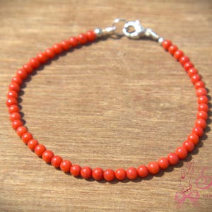 Puede incluir: Una delicada pulsera con pequeñas cuentas redondas de color rojo brillante. Se asegura con un cierre plateado. Las cuentas están espaciadas uniformemente, creando un diseño simple pero elegante. La pulsera se muestra sobre una superficie de madera.