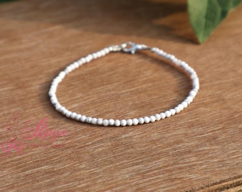 May include: A white beaded bracelet with a silver clasp lies on a wooden surface. The bracelet is made of small, round white beads. The background is a warm, natural wood grain. The bracelet is suitable for a wrist size of approximately 17 cm.