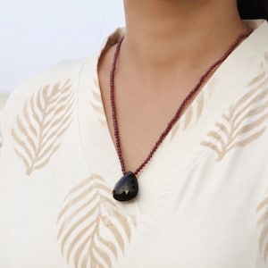 May include: A necklace featuring a dark, faceted, teardrop-shaped pendant suspended from a thin, beaded chain in a deep maroon color. The necklace is worn over a cream-colored top with a light brown leaf pattern printed across the fabric.
