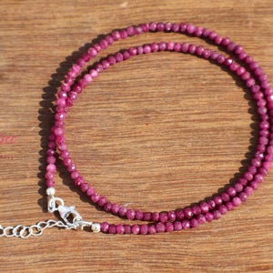 Puede incluir: Un delicado collar compuesto por pequeñas cuentas de rubí facetadas. El collar es de un color magenta vibrante y presenta un cierre y una cadena extensora de plata. La joya se exhibe sobre una superficie de madera.