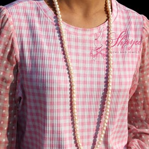 Pode incluir: Uma camisa rosa e branca xadrez de manga comprida com decote redondo. As mangas são transparentes com um padrão de bolinhas brancas. Um longo colar de pérolas pende na frente. A camisa tem o texto "Shayoo GEMS AND JEWELS".