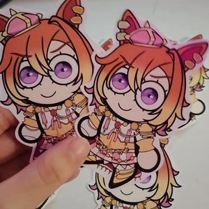 May include: A stack of stickers featuring a cartoon character with orange hair, purple eyes, and a pink and gold outfit. The character has cat-like ears and a crown. The stickers are cut around the character's outline.