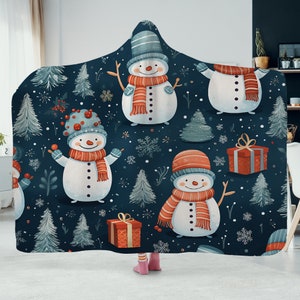May include: A hooded fleece blanket with a dark blue background and a repeating pattern of snowmen wearing red scarves and hats. The snowmen are surrounded by white snowflakes, green pine trees, and red gift boxes.