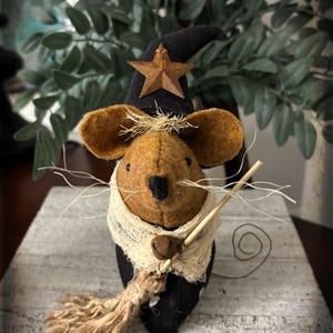 May include: A brown felt mouse figurine wearing a black witch hat adorned with a rusty metal star. The mouse has long white thread whiskers, a cream-colored fabric scarf with a small bell, and holds a miniature broom made of twine and wood.