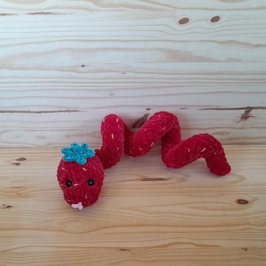 May include: A red crocheted snake toy with a blue flower on its head. The snake is curled up on a wooden surface.